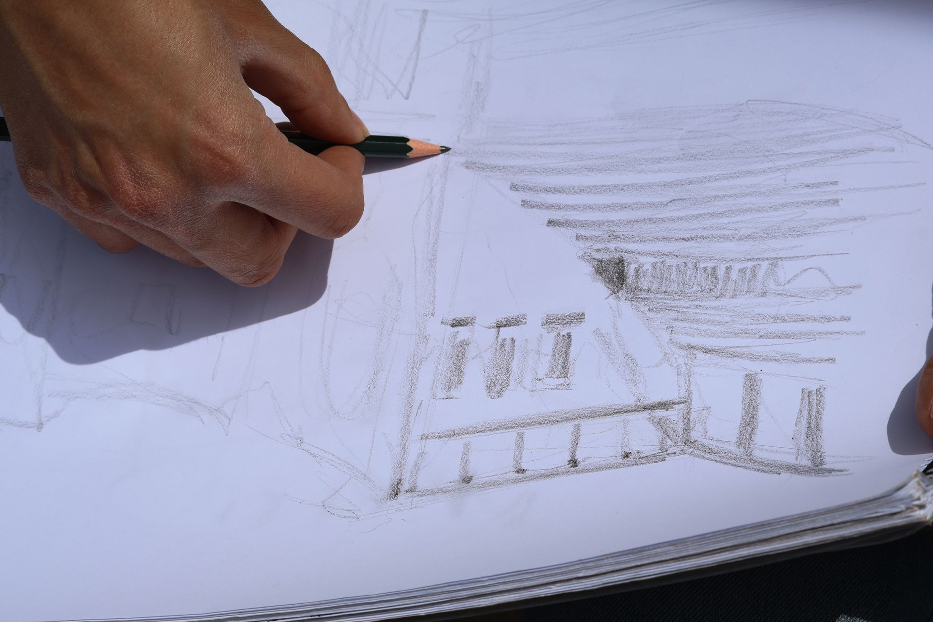 main tenant un crayon qui esquisse la façade d'un bâtiment dans un carnet, les grandes lignes posées avant tout détail