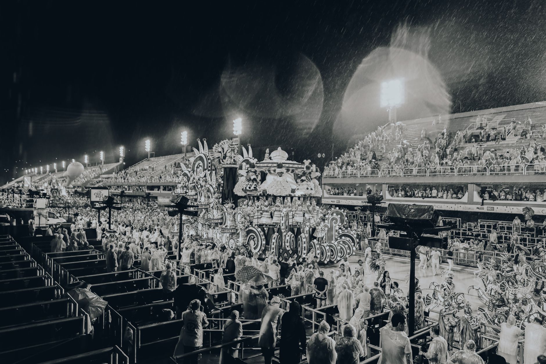 défilé nocturne sous une forte pluie au Sambódromo de Rio, char illuminé et spectateurs en ponchos transparents, photo en noir et blanc